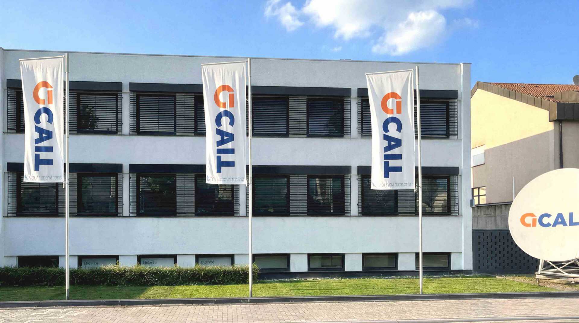 Firmengebäude der acall Technologies GmbH mit drei Fahnenmasten und acall-Logo-Bannern vor blauem Himmel.